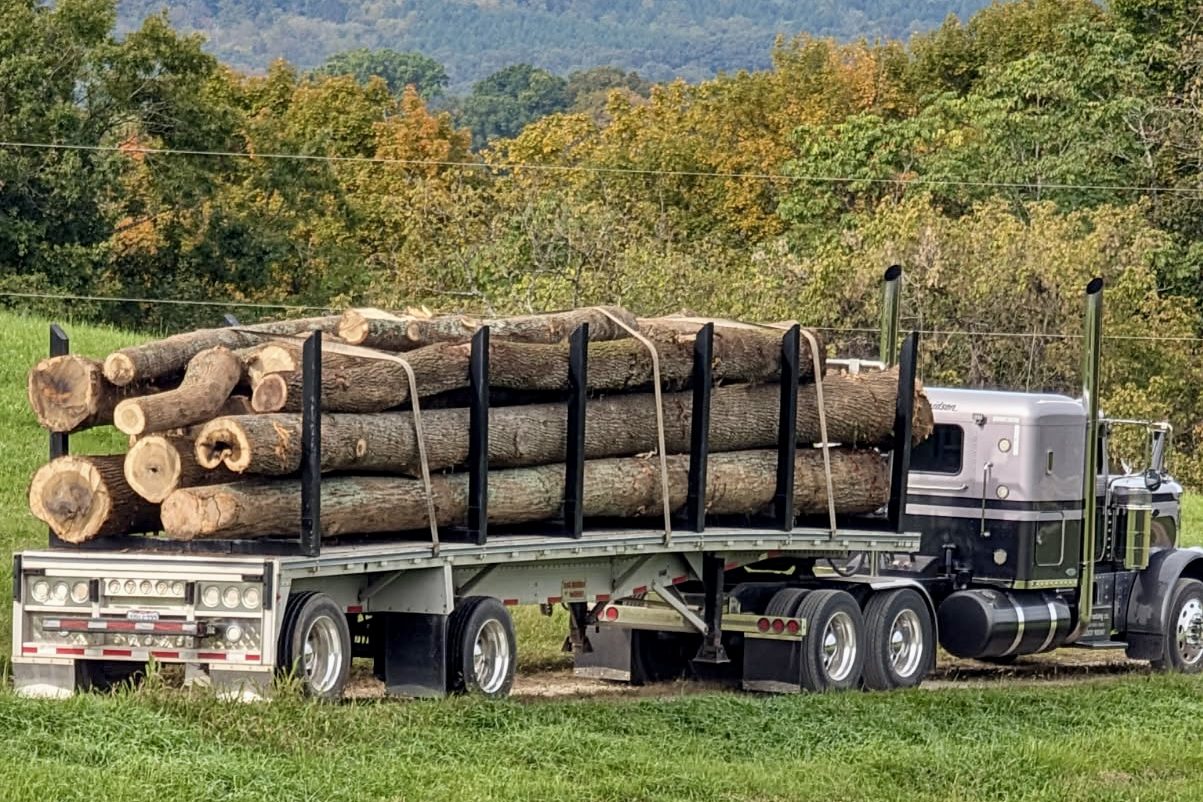A log truck loaded with hardwood logs at the edge of a field, wooded hills behind